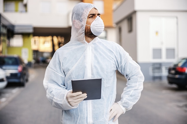 Homem com roupa de proteção contra vírus e máscara olhando e digitando no tablet, desinfetando edifícios de covid-19 com o pulverizador. prevenção de infecções e controle de epidemia. pandemia mundial.