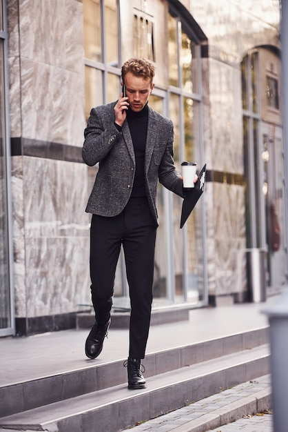 Homem com roupa formal elegante, com um copo de bebida e um bloco de notas nas mãos, está do lado de fora, contra um edifício moderno, tem uma conversa ao telefone.