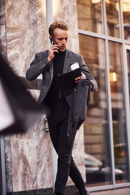 Homem com roupa formal elegante, com um copo de bebida e um bloco de notas nas mãos, está do lado de fora, contra um edifício moderno, tem uma conversa ao telefone.