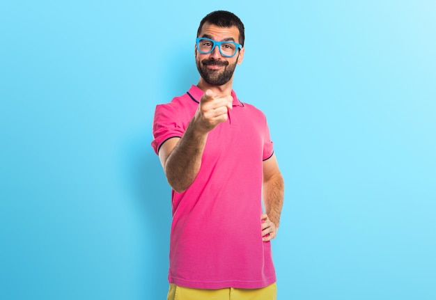 Homem com roupas coloridas apontando para a frente em fundo colorido