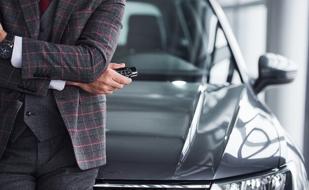 Homem com roupas formais modernas fica contra o carro preto com as chaves na mão.