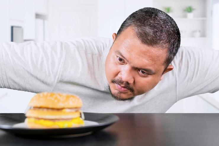 Foto homem com sobrepeso de meia-idade olhando para um cheeseburger pensando em comê-lo em casa