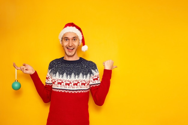 homem com suéter de Natal com chapéu de Papai Noel vermelho de rena aponta o dedo para uma área vazia