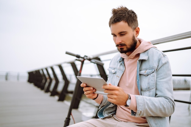 Homem com tablet andando de scooter elétrica no cais perto do mar Freelancer Transporte ecológico na cidade