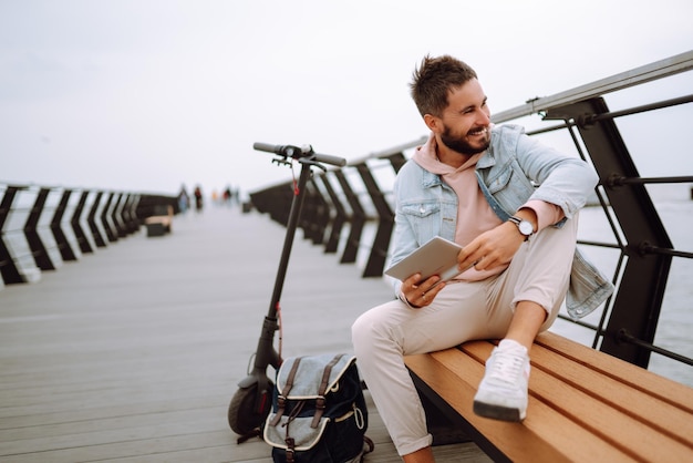 Homem com tablet andando de scooter elétrica no cais perto do mar Freelancer Transporte ecológico na cidade