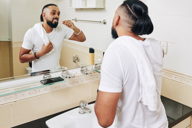 Foto homem com toalha no ombro, escovando os dentes em frente ao espelho do banheiro pela manhã