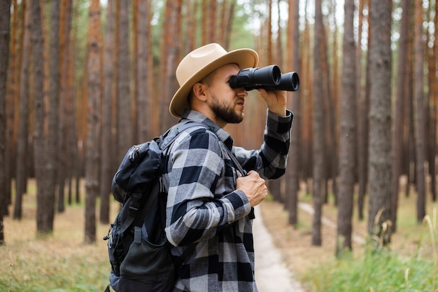 Homem com um chapéu e uma camisa xadrez segura binóculos e caminha pela floresta.