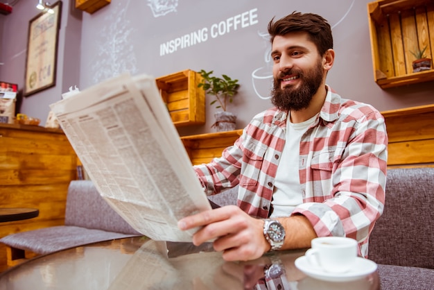 Homem com uma barba que lê um jornal e que bebe o café.