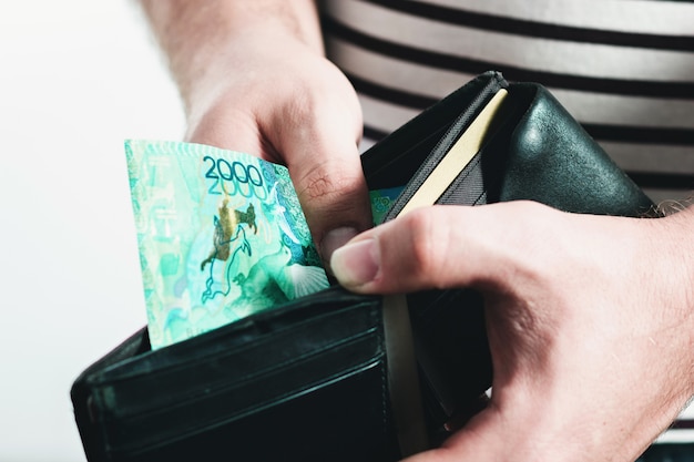 Homem com uma camiseta listrada, segurando uma carteira de couro preto com notas do cazaquistão.