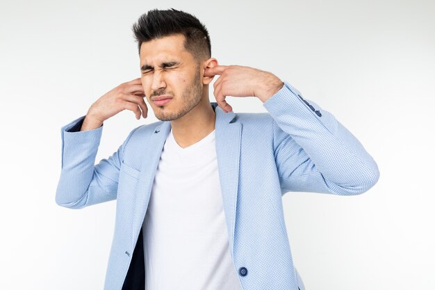 Homem com uma jaqueta clássica azul tapa os ouvidos com os dedos contra barulho alto em um fundo branco.