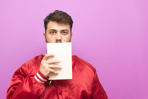 Homem com uma jaqueta vermelha fica em um fundo rosa com um livro branco