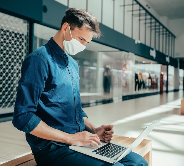 Homem com uma máscara protetora funciona em um laptop em um prédio de shopping center.