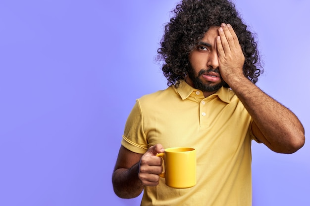 Homem com uma xícara de chá, homem árabe encaracolado precisa dormir, fechando um olho, posando para a câmera isolada no espaço roxo