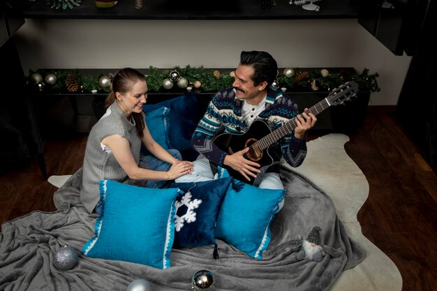 Homem com violão cantando para a namorada sentado na sala de estar com almofadas em uma noite de Natal