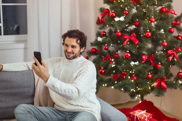 Homem comemorando ano novo perto da árvore de Natal em videochamada doméstica ou selfie no smartphone