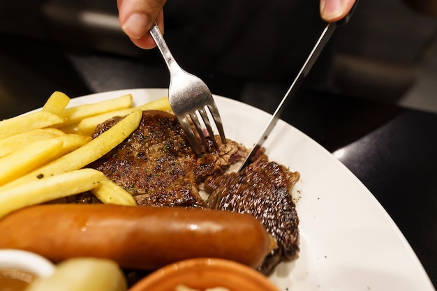 Homem comendo estaca de carnes grelhadas do prato mão segurando faca e garfo cortando bife grelhado