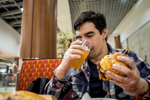 Homem comendo hambúrguer de fast food e bebendo cerveja sozinho na área aberta de um restaurante em um shopping center.