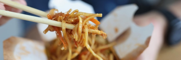 Homem comendo macarrão chinês com pauzinhos fora da caixa de papel closeup