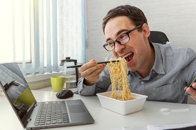 Homem comendo macarrão instantâneo enquanto trabalha com o laptop no escritório