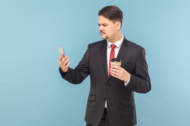 Homem concentrado sério com bigode em pé usando telefone celular e bebendo café para viagem
