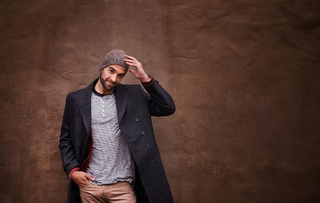 Foto homem confiança e sorriso com moda urbana e elegante casaco de cavalheiro modelo inverno e boné com felicidade da cidade ou positividade ao ar livre para estilo hipster legal isolado em fundo de parede marrom