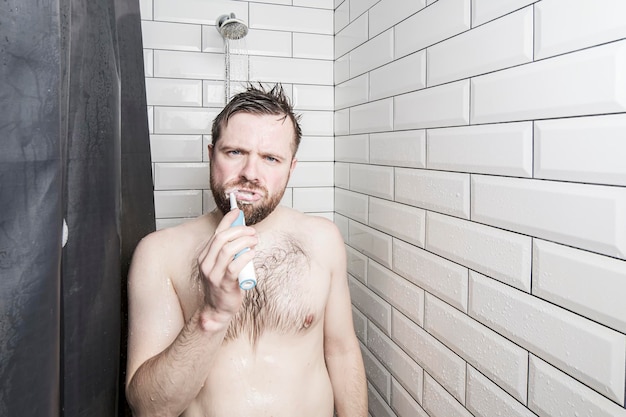 Homem confiante escova os dentes com uma escova de dentes recarregável em pé no chuveiro sob água corrente