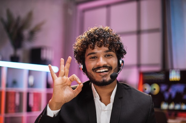 Homem confiante positivo sorridente em roupa formal e fone de ouvido sentado na mesa do escritório