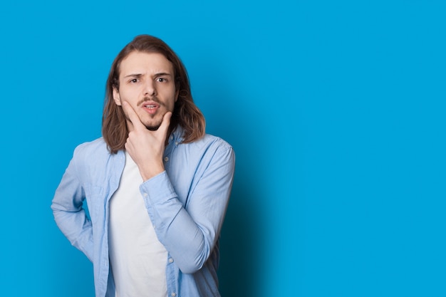 Homem confuso com cabelo comprido e cerdas tocando o queixo com a mão posando em roupas casuais em um azul