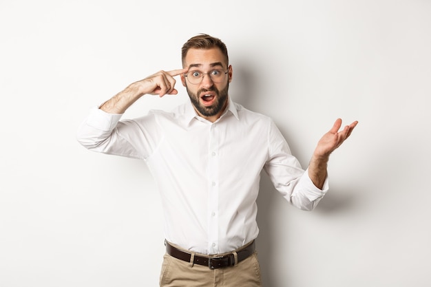 Homem confuso e chocado, apontando para a cabeça, repreendendo o funcionário por agir estúpido, em pé sobre um fundo branco.