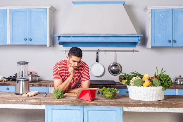 Homem confuso pensativo assistindo blog de comida usando tablet na cozinha moderna procurando receita de prato na internet para cozinhar salada vegetariana com vegetais frescos verdes nutrição saudável dieta vegana