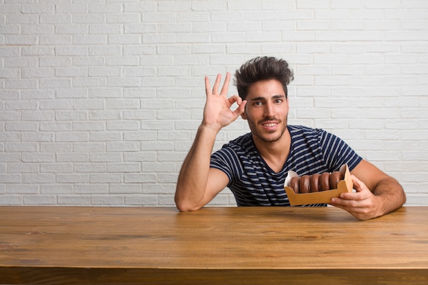 Homem considerável e natural novo que senta-se em uma tabela alegre e seguro fazendo o gesto aprovado, entusiasmado e gritando, conceito da aprovação e sucesso. Comendo rosquinhas de chocolate.