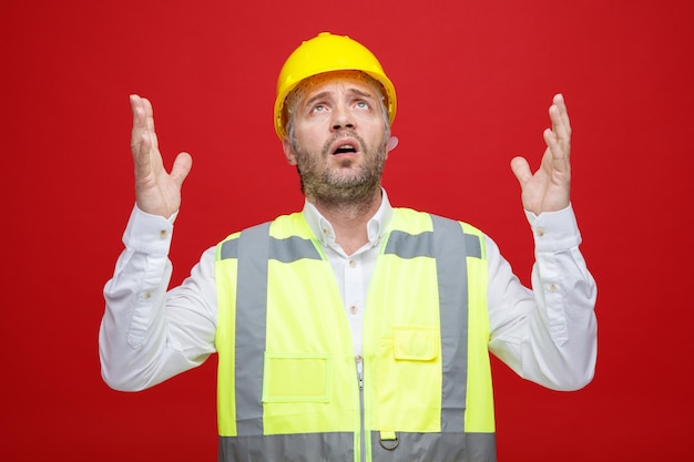 Homem construtor em uniforme de construção e capacete de segurança olhando confuso e preocupado levantando os braços sobre fundo vermelho