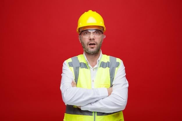 Homem construtor em uniforme de construção e capacete de segurança olhando para a câmera espantado e surpreso com os braços cruzados no peito em pé sobre fundo rosa