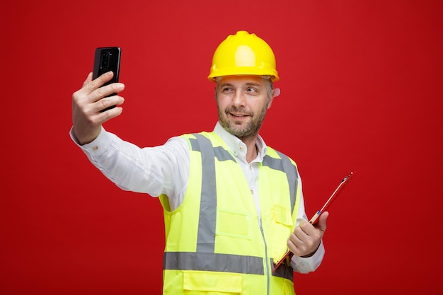 Homem construtor em uniforme de construção e capacete de segurança segurando a área de transferência fazendo selfie usando smartphone sorrindo confiante em pé feliz e positivo sobre fundo vermelho