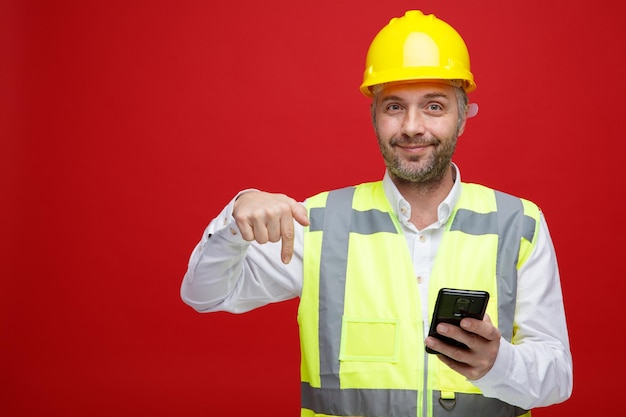 Homem construtor em uniforme de construção e capacete de segurança segurando o smartphone apontando com o dedo indicador para baixo olhando para a câmera sorrindo em pé sobre fundo vermelho