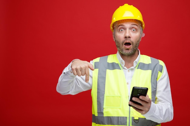 Homem construtor em uniforme de construção e capacete de segurança segurando o smartphone apontando com o dedo indicador para baixo olhando para a câmera surpreso em pé sobre fundo vermelho