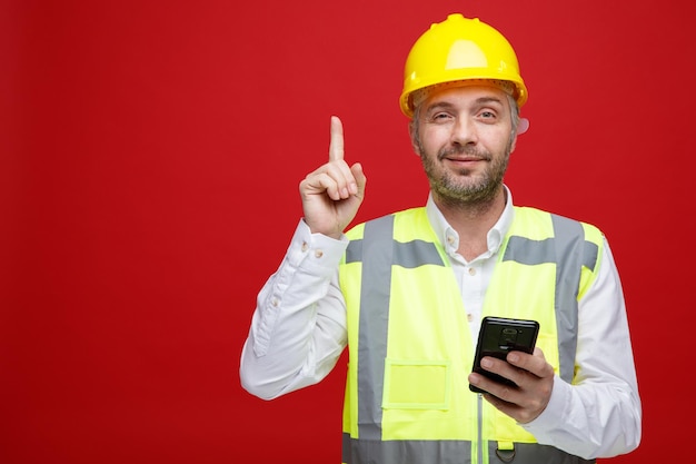 Homem construtor em uniforme de construção e capacete de segurança segurando smartphone olhando para câmera sorrindo confiante mostrando o dedo indicador sorrindo em pé sobre fundo vermelho