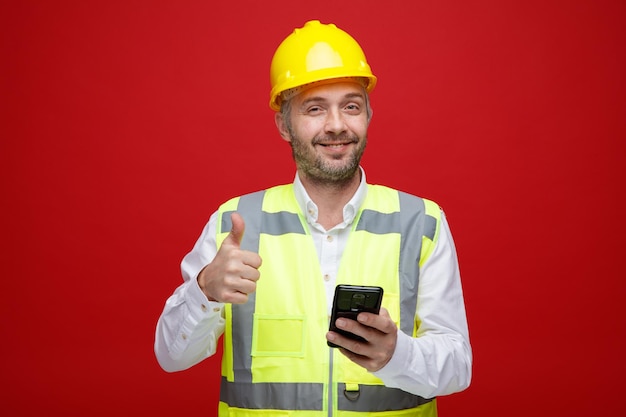 Homem construtor em uniforme de construção e capacete de segurança segurando smartphone olhando para câmera sorrindo mostrando o polegar em pé sobre fundo vermelho