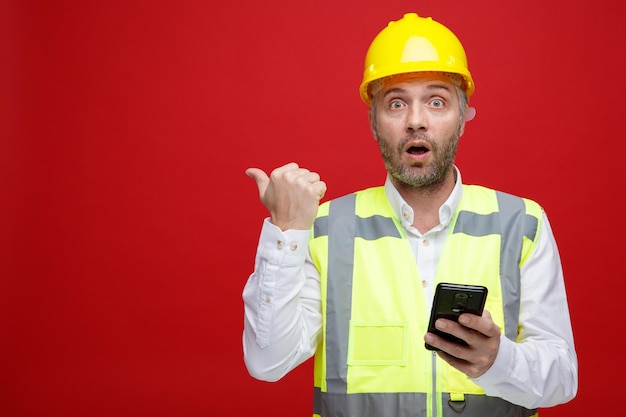 Homem construtor em uniforme de construção e capacete de segurança segurando smartphone olhando para câmera surpreso apontando com o polegar para o lado em pé sobre fundo vermelho