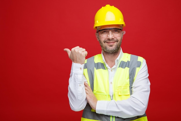 Homem construtor em uniforme de construção e capacete de segurança usando óculos de segurança, olhando para a câmera com sorriso no rosto, apontando com o polegar para o lado em pé sobre fundo rosa