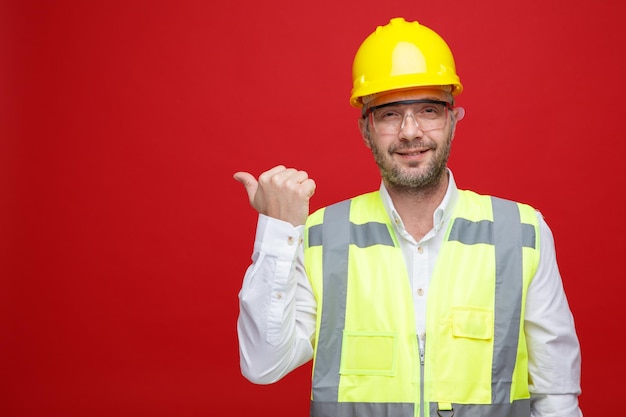 Homem construtor em uniforme de construção e capacete de segurança usando óculos de segurança, olhando para a câmera com um sorriso no rosto, apontando com o polegar para o lado em pé sobre fundo rosa