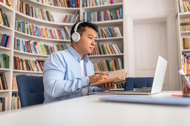 Homem coreano aprendendo online segurando livro usando laptop na biblioteca