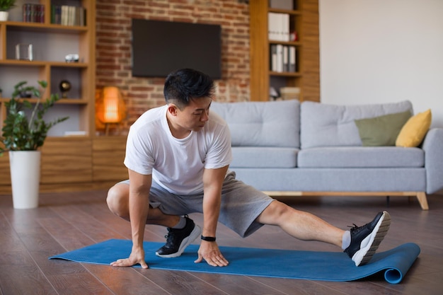 Homem coreano de treino em casa esticando as pernas no tapete de ioga na sala de estar com espaço livre para treino doméstico