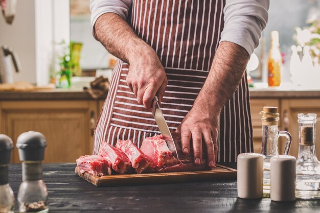 Homem corta um pedaço de carne fresca em uma tábua de madeira na cozinha de casa. Um homem com um avental listrado com uma grande faca nas mãos