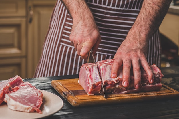 Foto homem corta um pedaço de carne fresca em uma tábua de madeira na cozinha de casa. um homem com um avental listrado com uma grande faca nas mãos