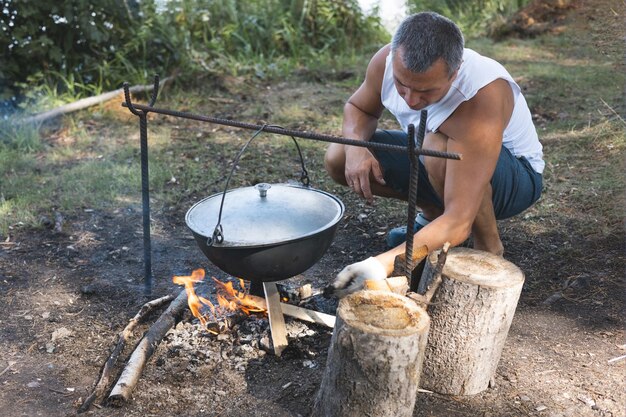 Homem cozinhando no caldeirão em fogo aberto na natureza Panela na fogueira no fundo da floresta Vida de acampamento Viagens locais