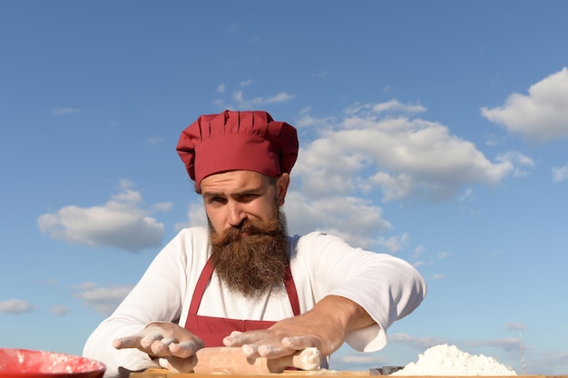 Homem cozinheiro chef com barba no rosto bonito em uniforme branco e vermelho cozinhar massa ensolarada ao ar livre no fundo do céu azul