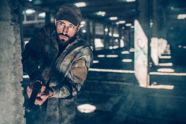 Homem cuidadoso está olhando para a frente. Ele está de pé atrás da coluna em um grande hangar. Warrior ha uma pequena arma preta. Ele está pronto para filmar a qualquer momento.