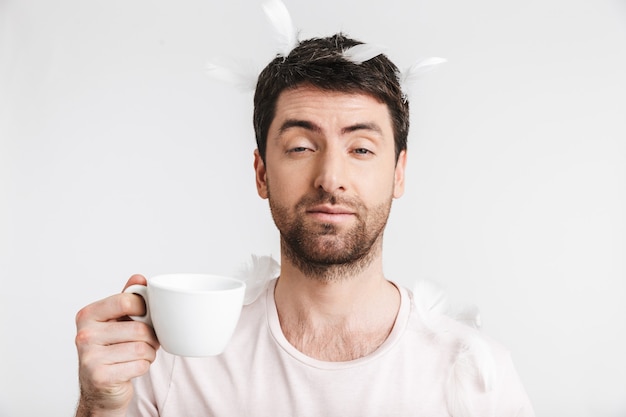 Homem da manhã com cerdas em uma camiseta casual bebendo café enquanto fica de pé sob as penas que caem, isoladas sobre uma parede branca