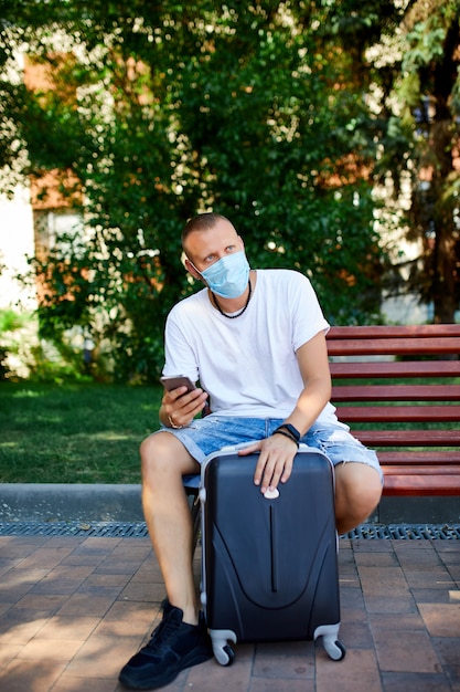 Homem da máscara protetora, sentado no parque ao ar livre com uma mala e um telefone celular